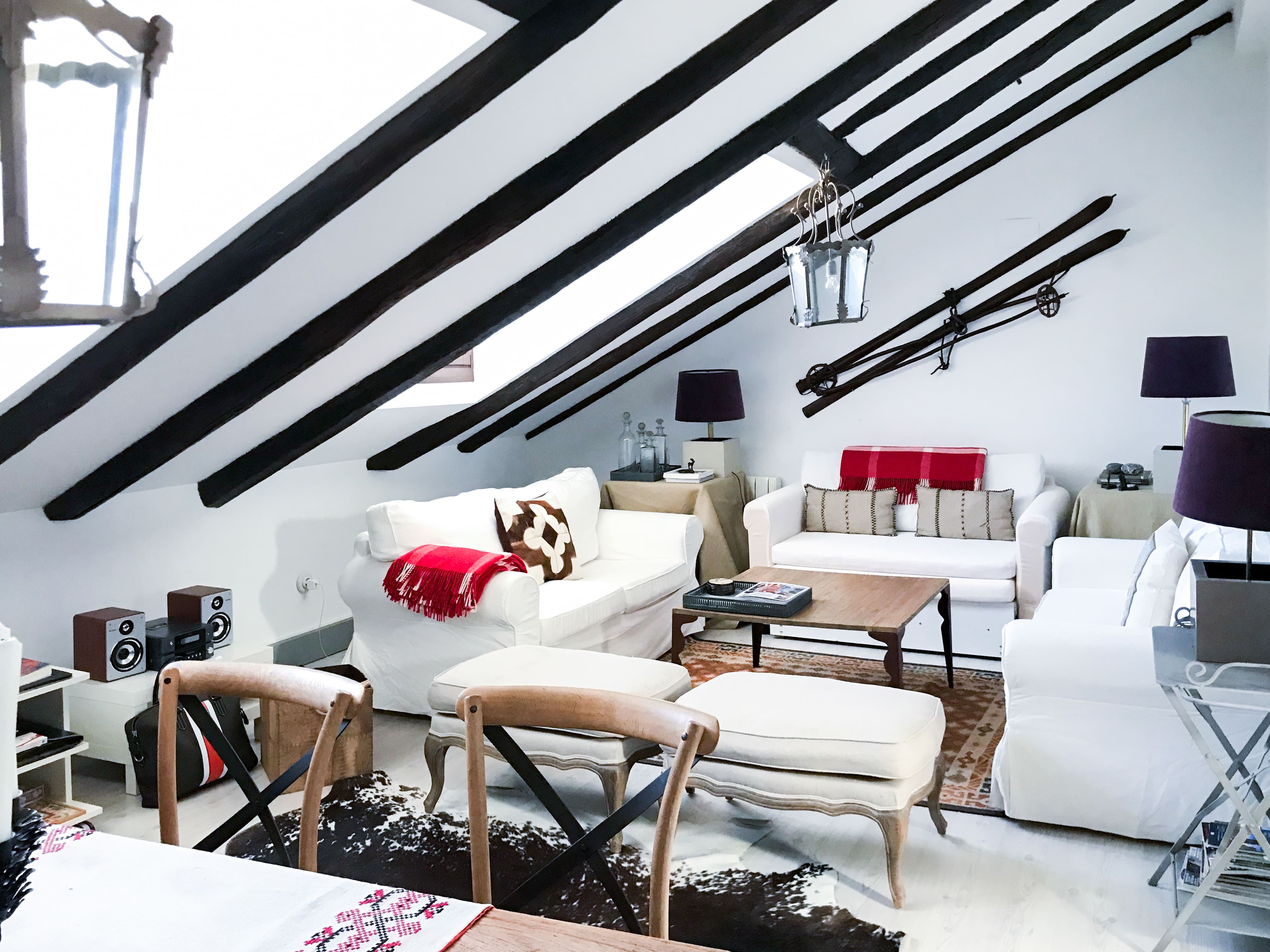 Salón abuhardillado con vigas negras, sofás blancos y esquís antiguos en la pared