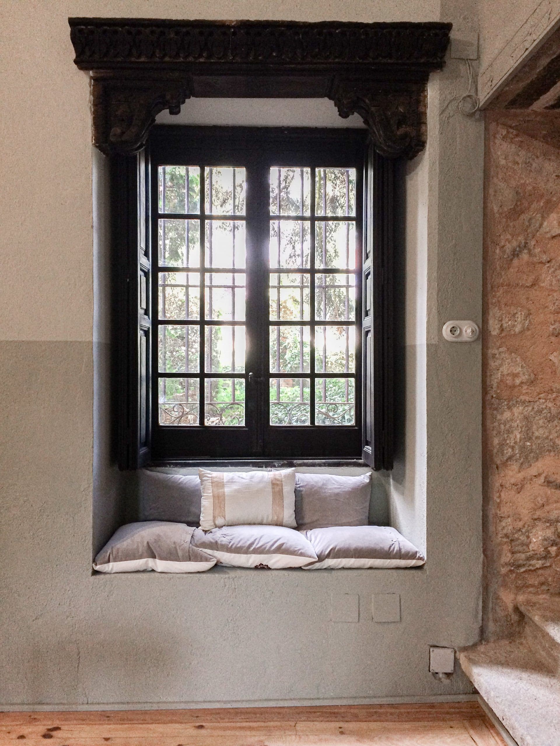 Ventanal de carpintería negra en muro de piedra, con asiento de obra y cojines de lino