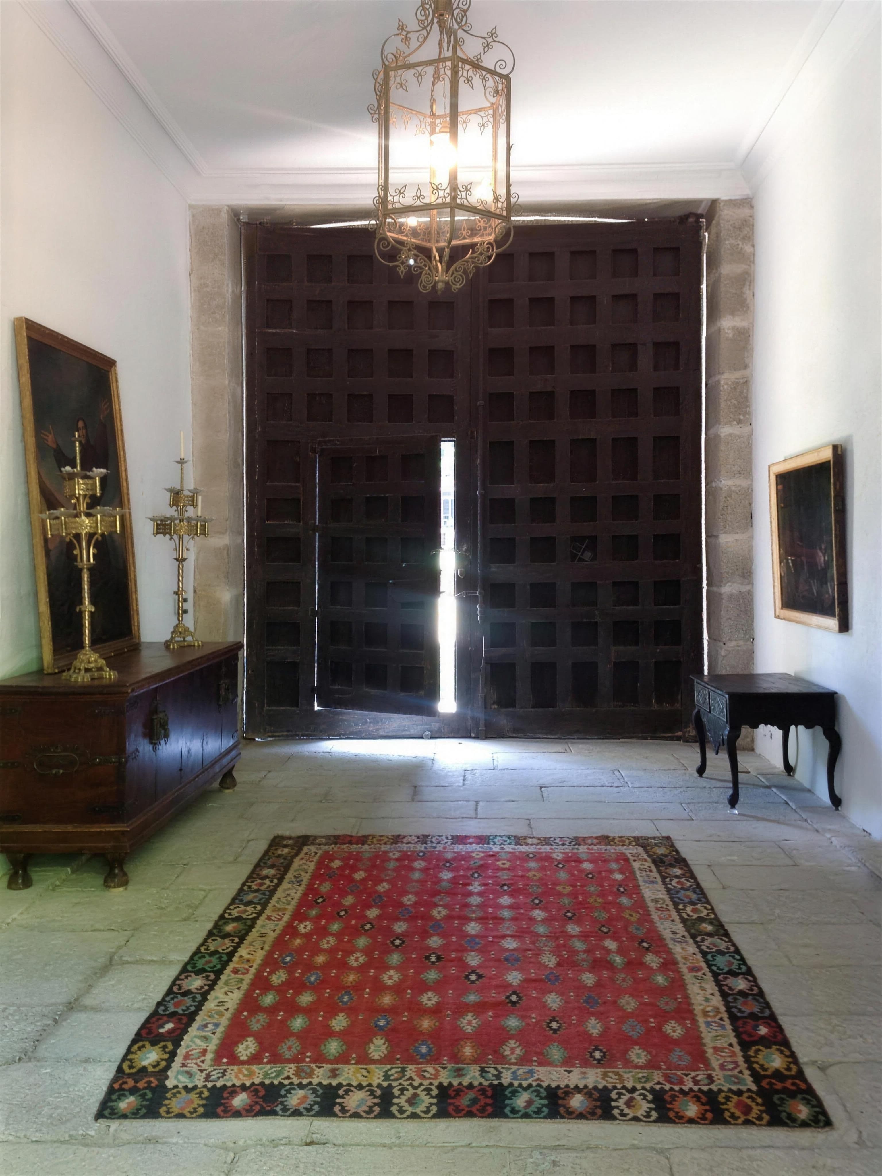 Portón histórico de madera claveteada entreabierto, con kilim rojo y farol de bronce