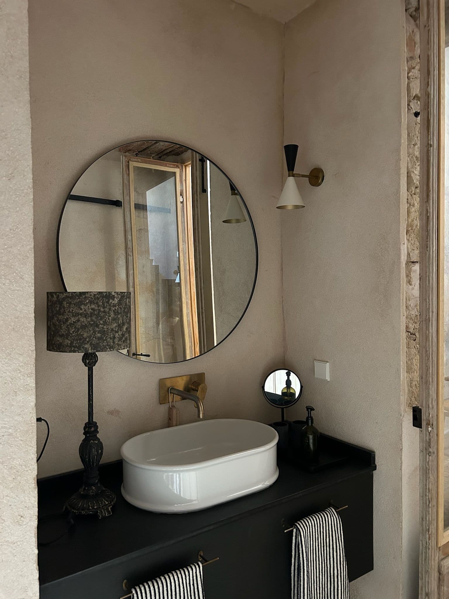 Baño con espejo redondo, lavabo blanco sobre mueble negro y grifería de latón