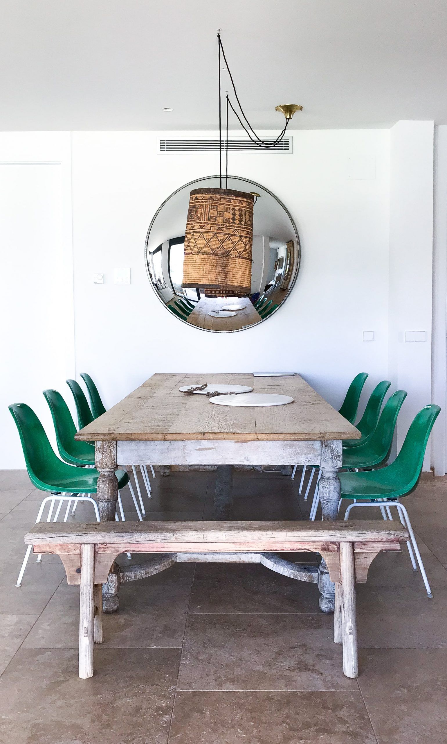 Comedor con gran mesa de madera decapada, sillas verdes y espejo convexo bajo lámpara de fibra