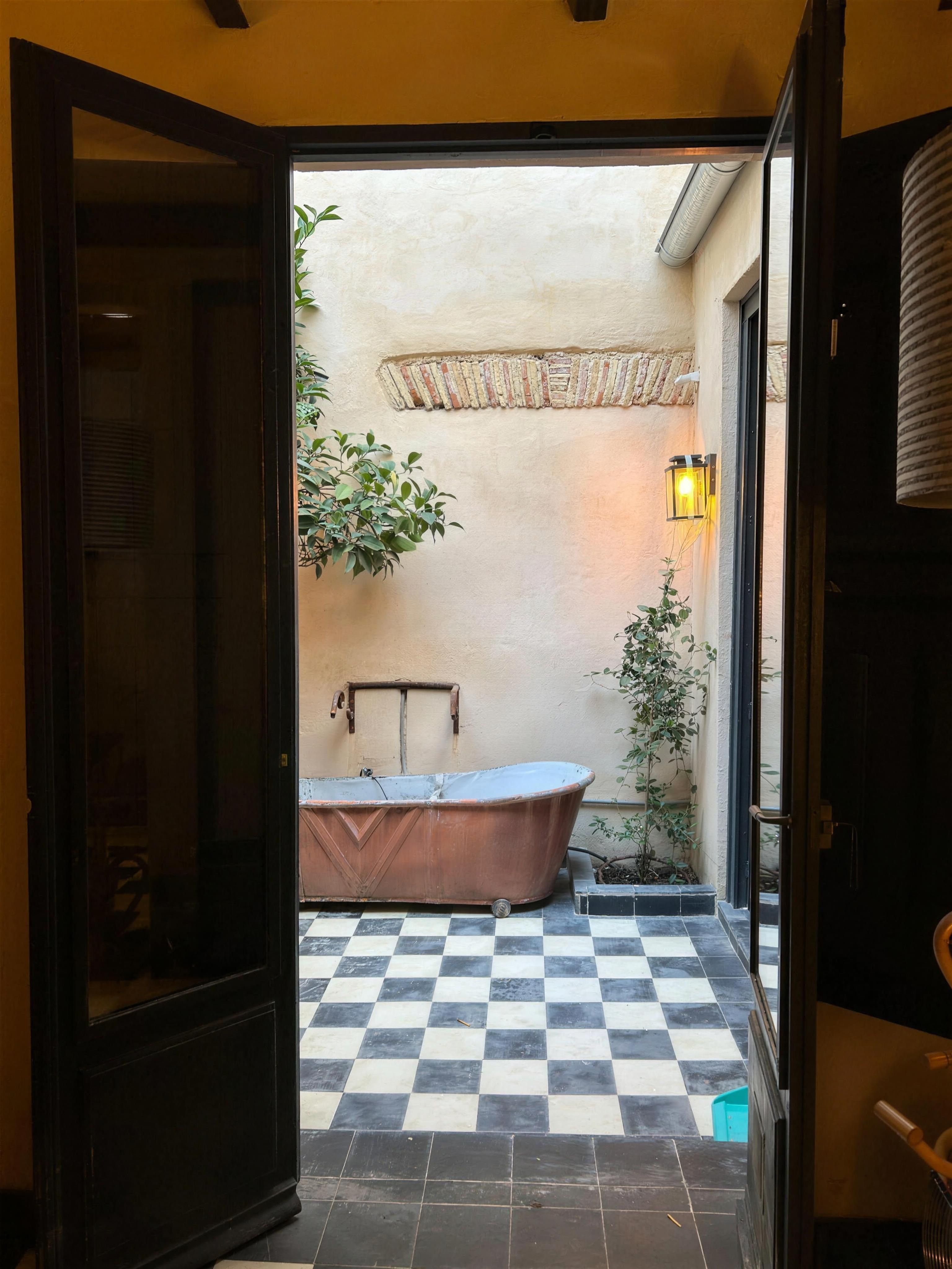 Patio interior al anochecer con bañera exenta de cobre, suelo damero en blanco y negro y un cítrico en maceta