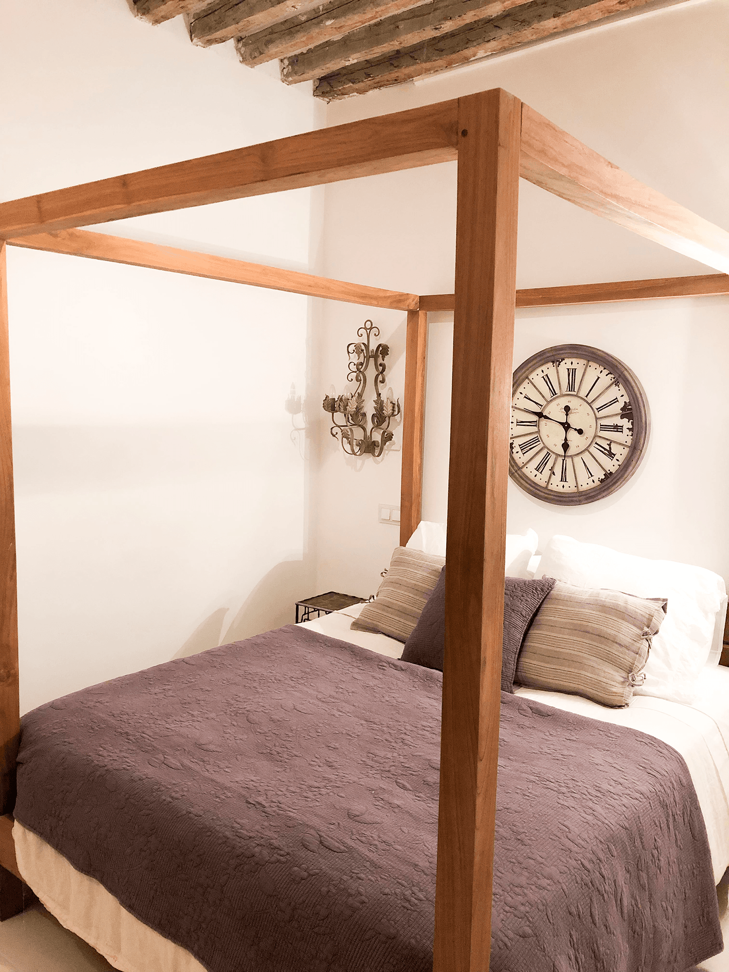 Cama con dosel de madera clara, colcha morada y reloj de estación en la pared