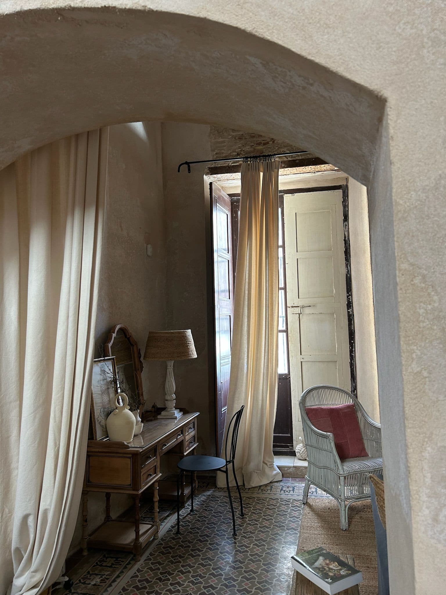 Arco encalado con cortinas de lino hacia el salón, cómoda antigua y butaca de mimbre