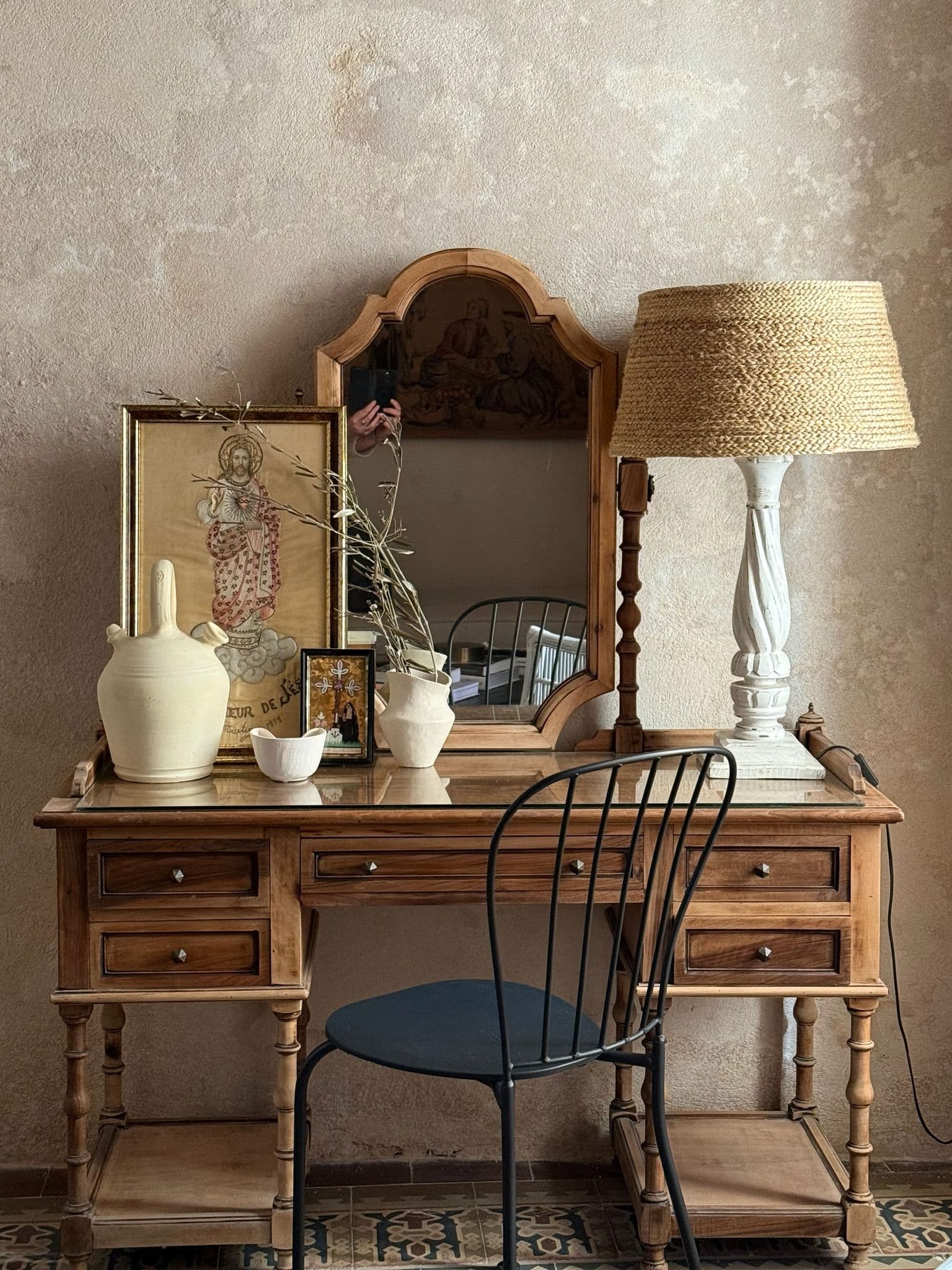 Escritorio antiguo de madera con lámpara de rafia, silla de hierro y estampas sobre muro encalado