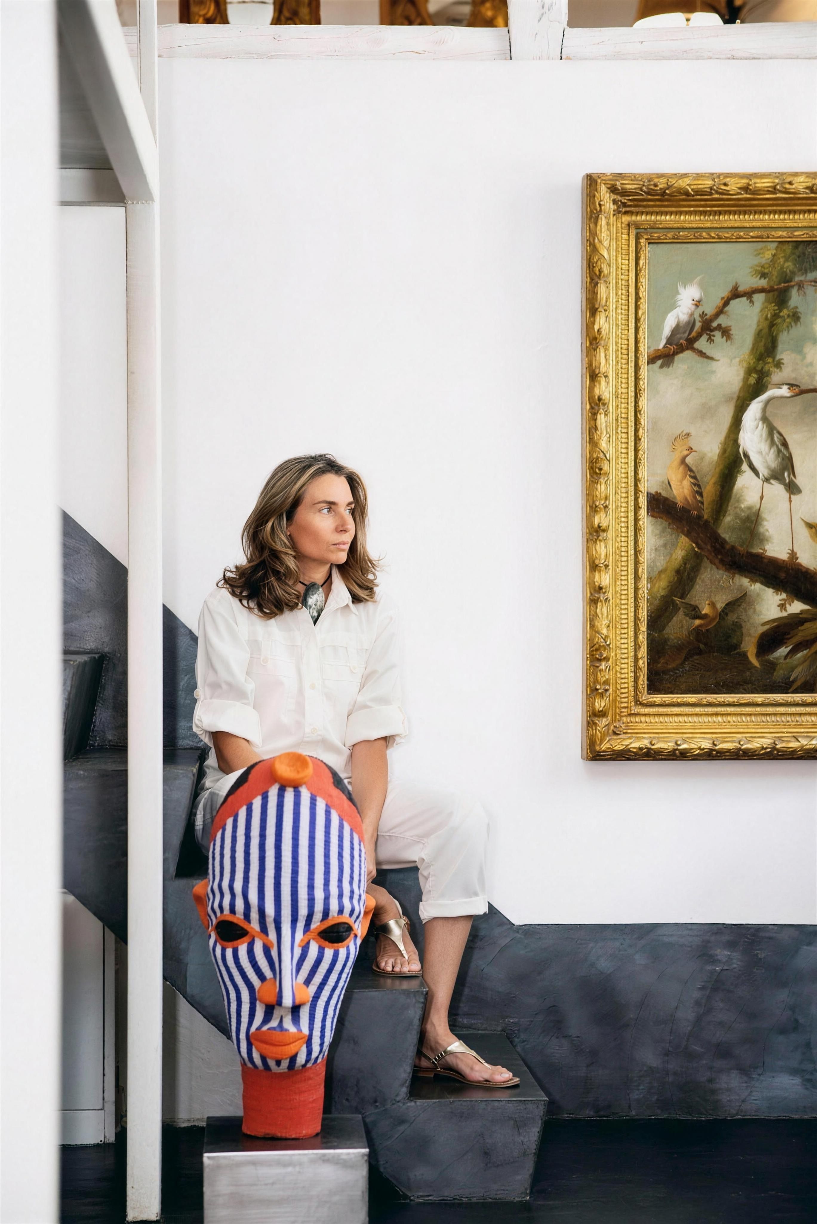Rebeca Ruiz sentada en una escalera grafito, de blanco, junto a una máscara africana de rayas y un óleo clásico de pájaros con marco dorado