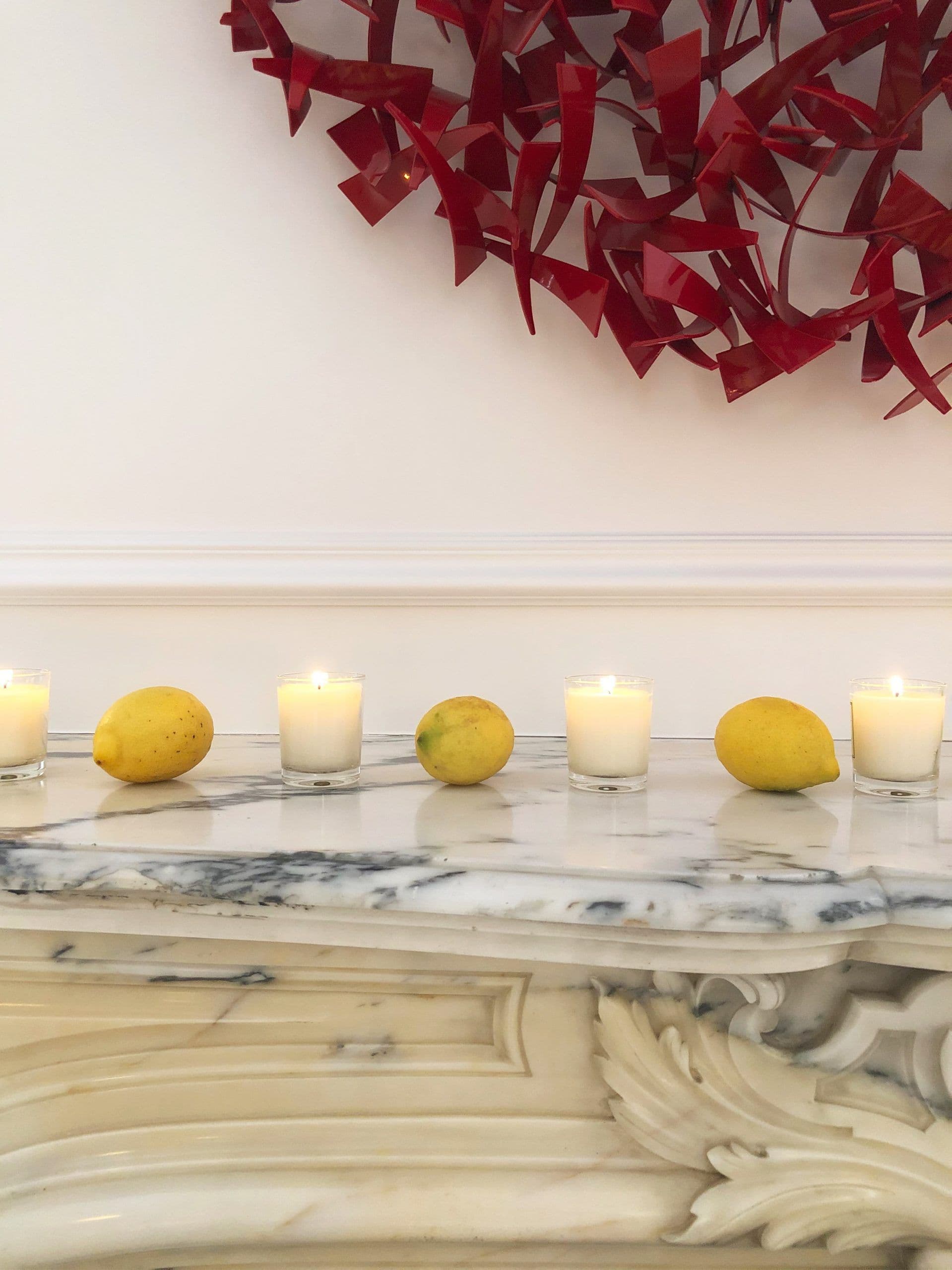 Limones y velas sobre repisa de mármol tallado, con escultura roja de lazos en la pared